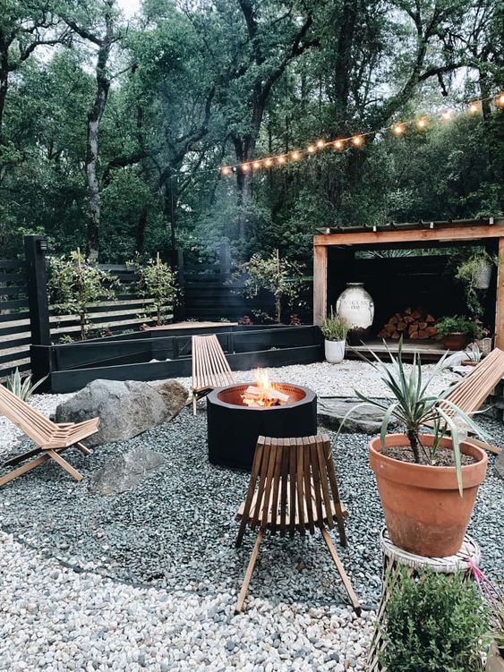 a lovely modern gravel patio with a black fire pit, stained wooden chairs, potted greenery and large rocks