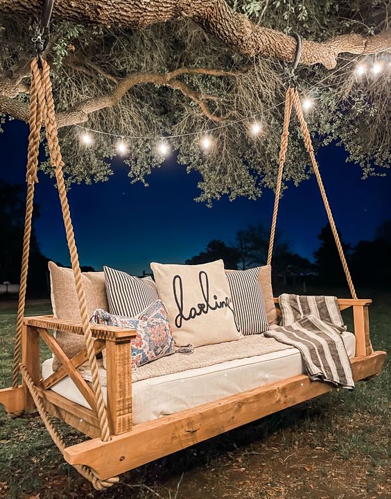 a lovely outdoor swinging daybed in rustic style,w ith lots of pillows and lights over it is a cool idea