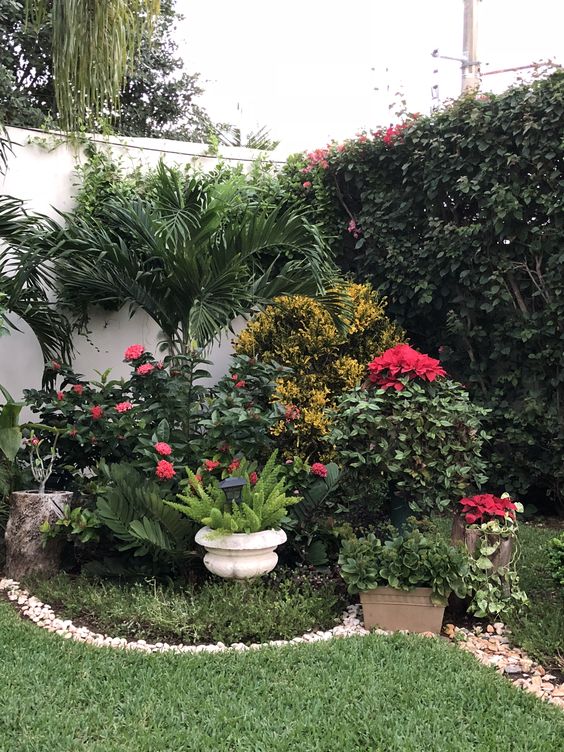 a lush corner garden with grasses, potted greenery and blooms, some trees, a living wall and pebbles covering the border of the flower bed