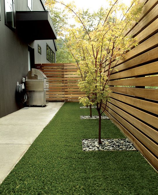 a minimal side yard with a light-stained wooden fence, a green lawn, some trees and a grill in the corner