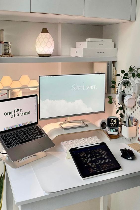 a minimal white workspace with a shelf with decor and lamps, a memo board with devices and stuff and some monitors on the desk