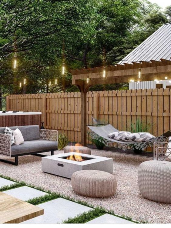 a modern outdoor area with a hammock, grey chairs, poufs, a fire pit, some greenery and lights over the space