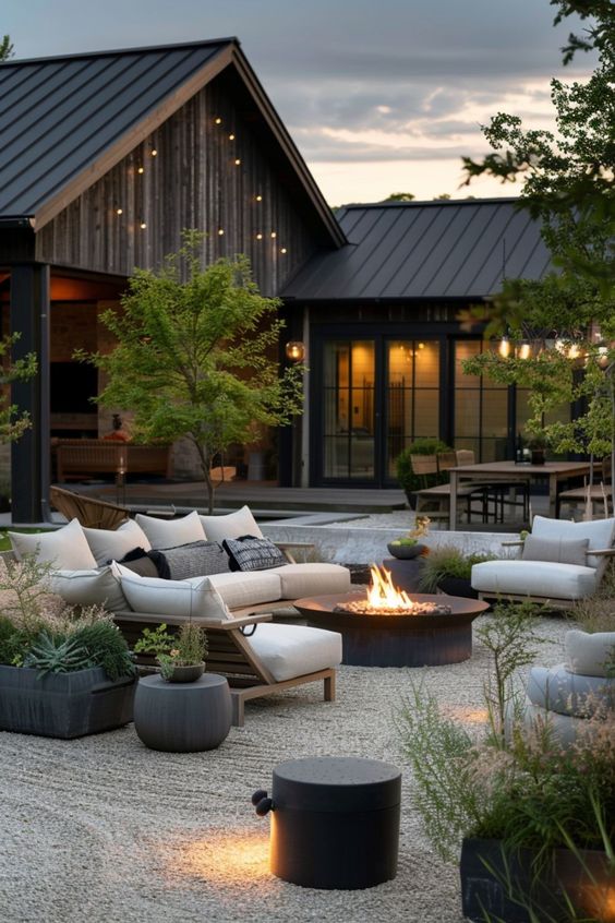 a modern and chic gravel patio with a metal fire pit, white seating furniture with pillows, potted plants around and some modern lights