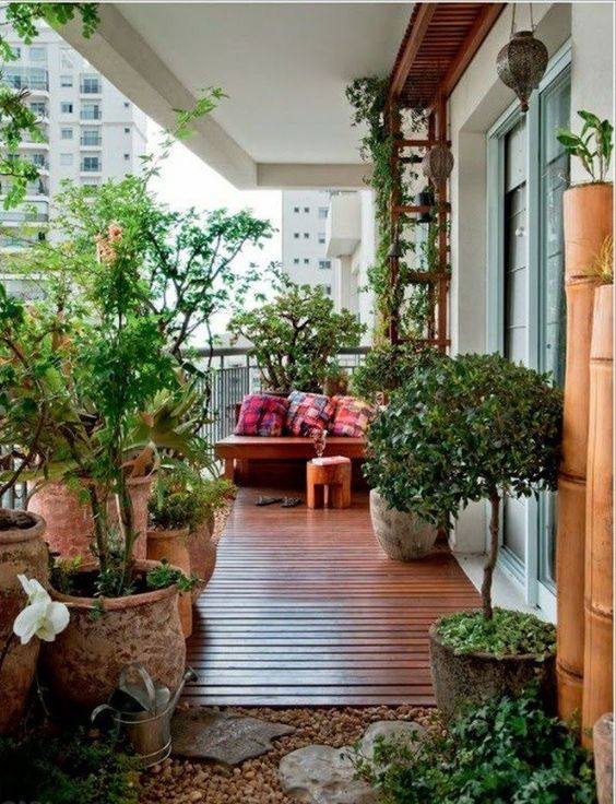 a modern balcony with a planked floor and some rocks, potted plants, a corner sofa with pillows, a trellis with greenery