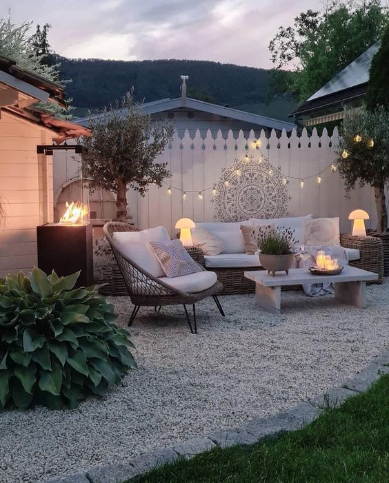 a modern boho patio with wicker seating furniture, a fire pit, a boho chair, some trees, lights and decor