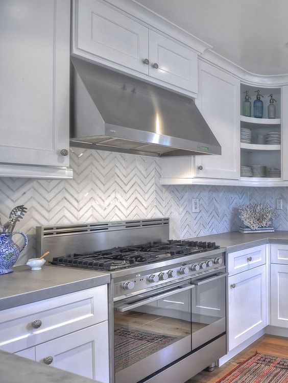 a modern farmhouse white kitchen with grey stone countertops and a glossy chevron tile backsplash plus stainless steel appliances