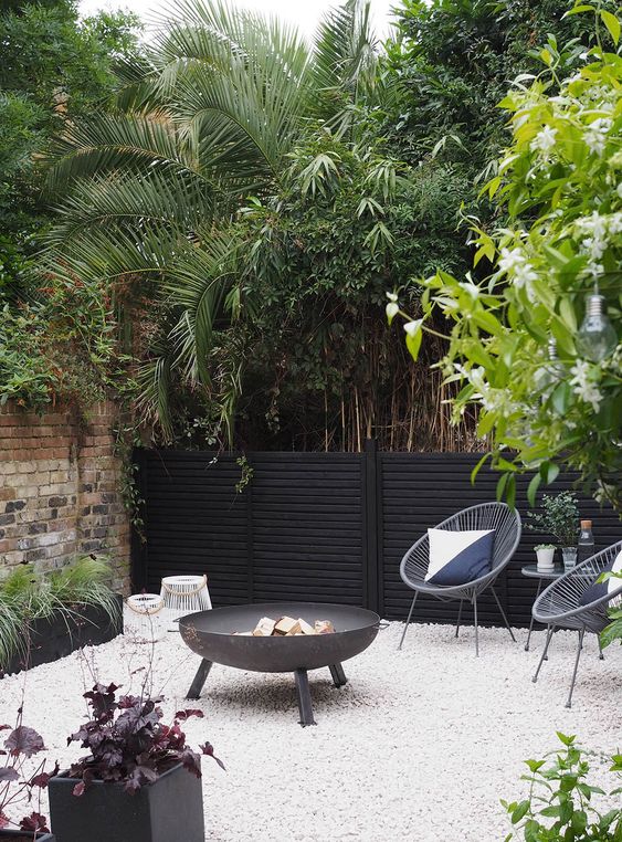 a modern gravel patio with a black fire pit, grey chairs with pillows, candle lanterns, potted greenery and trees around for a modern space