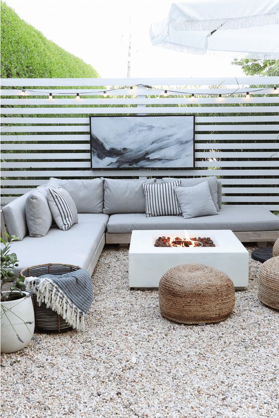 a modern gravel patio with a corner sofa, a fire pit, jute poufs, lights and an umbrella and potted plants around