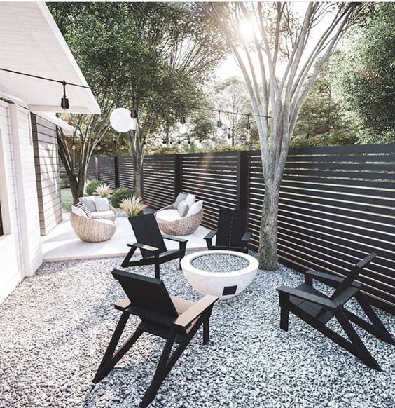 a modern gravel patio with a fire pit, black chairs, a deck with wooden seating furniture and lights over the space