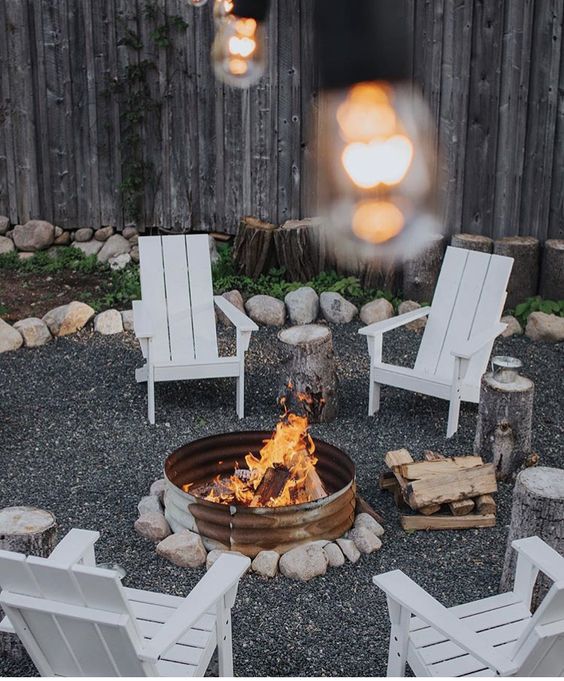 a modern gravel patio with a metal fire pit, white chairs, rocks around and stumps as side tables is cozy