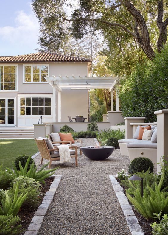 a modern gravel patio with a sofa with pillows, a fire pit, chairs with pillows, planted greenery around