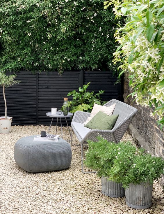 a modern gravel patio with wicker seating furniture, a pouf, planters with greenery and a side table with greenery