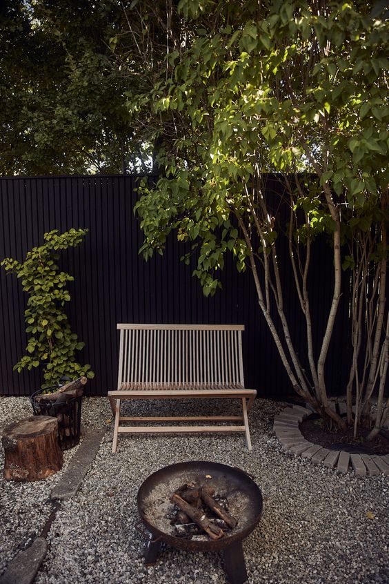 a modern gravel patoi with a wooden benchm tree stumps, a metal fire pit, trees and a black fence