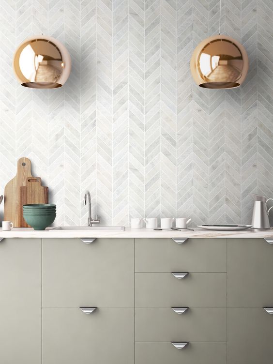 a modern kitchen with sage grene cabinets, white countertops and a grey marble chevron backsplash plus gold pendant lamps