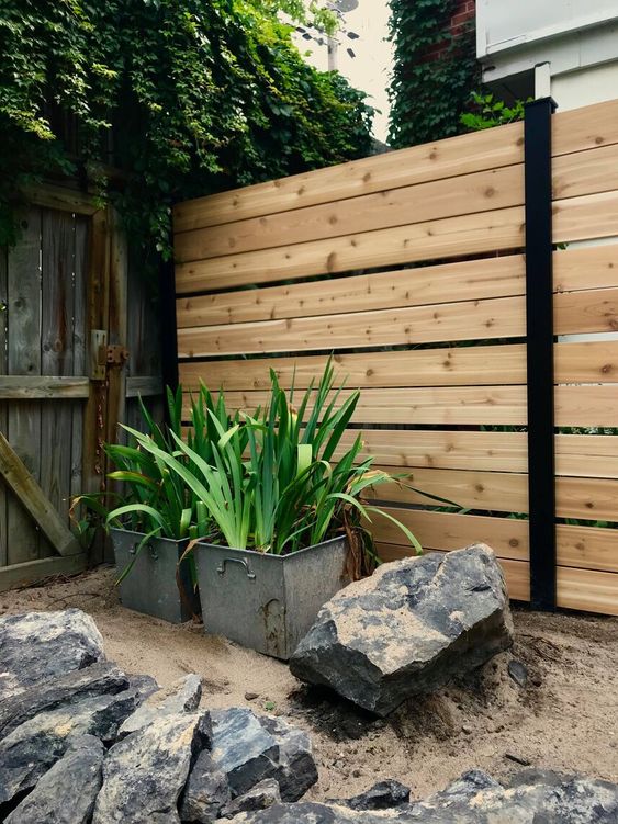 a modern light-stained fence with blackened metal touches, potted greenery and oversized rocks are a chic combo for a modern backyard