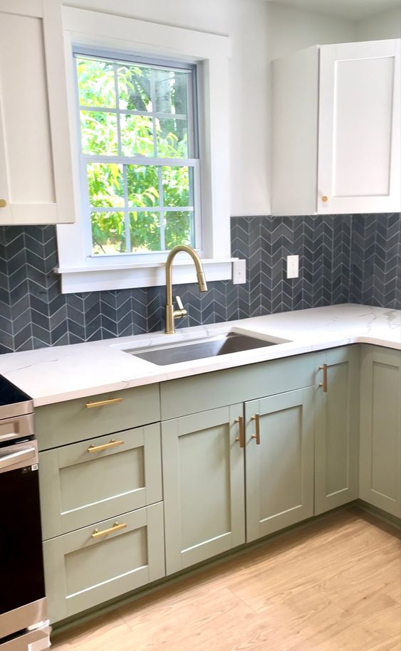 a modern sage green and white kitchen with a grey chevron tile backsplash and white countertops plus gold fixtures