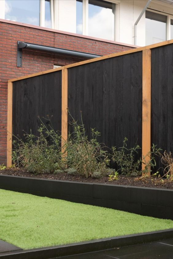 a modern tall wooden fence with black wood and light-stained frames, a long flower bed with greenery for a fresh feel