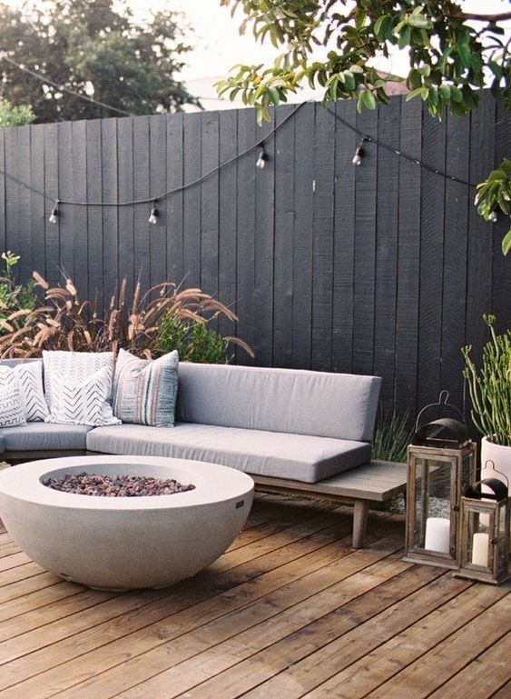 a modern terrace with a corner sofa, a fire pit, candle lanterns, greenery and a black wooden fence with lights