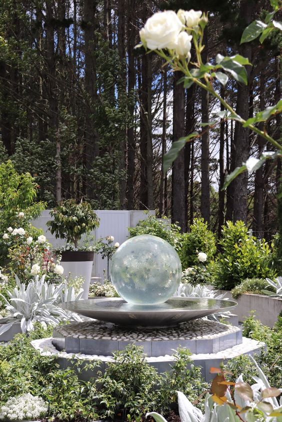 a modern white garden with greenery and white blooms around, a stand with a water bowl and a glass ball is a lovely idea for any space