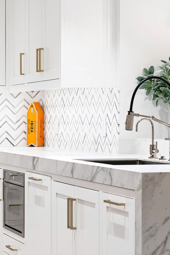 a modern white kitchen with shaker cabinets, a chevron tile backsplash and white marble countertops plus greenery