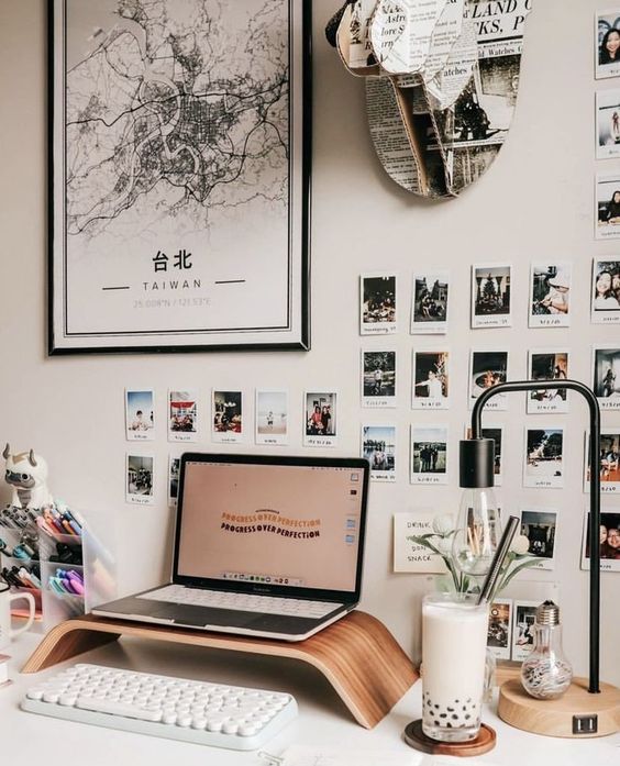 a modern workspace with a laptop on a stand, a keyboard, a gallery wall with Polaroids, some maps and cool accessories