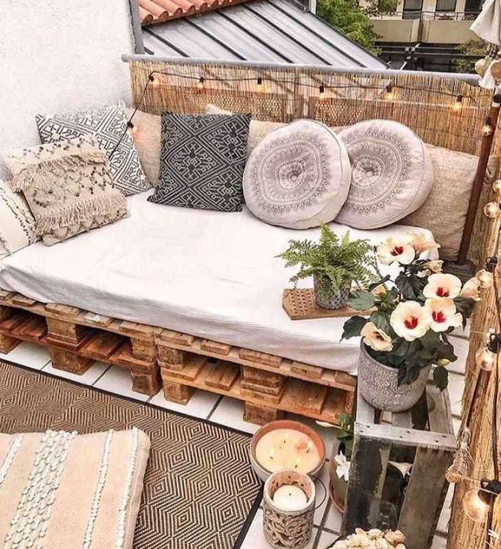 a neutral boho balcony with a pallet daybed, neutral pillows with tassels, potted plants and blooms, candles in candleholders and layered rugs