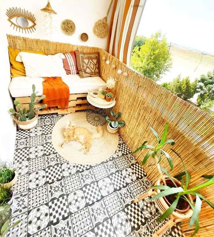 a pretty boho balcony with black and white tiles on the floor, a pallet seat with pillows, potted greenery, a jute rug and some baskets and lamps