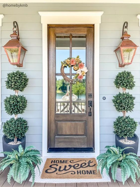 a pretty farmhouse porch with a stained door and a wreath, tall topiary trees, copper lanterns and potted greenery