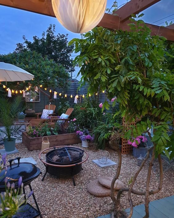 a pretty gravel patio with a fire pit, a grill, wooden loungers, a raised garden bed with blooms, lights, trees and a trellis is amazing