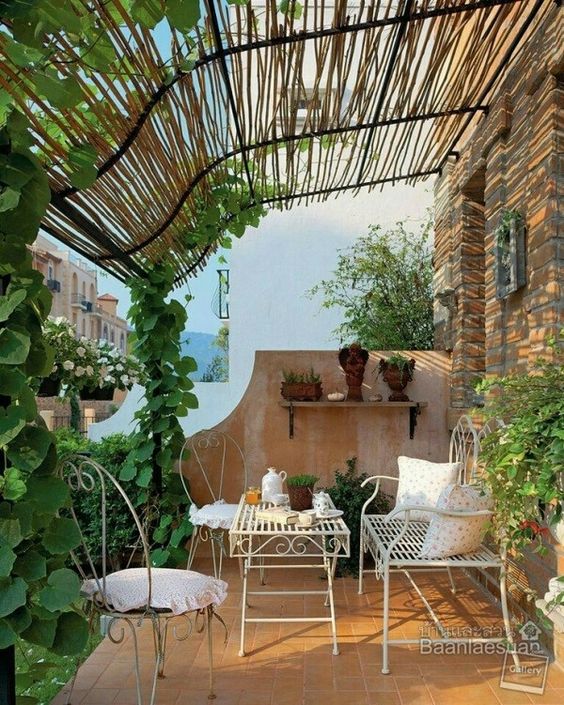 a Provence inspired balcony with white forged furniture, potted greenery and blooms and a trellis with vines climbing up the pillars to the roof