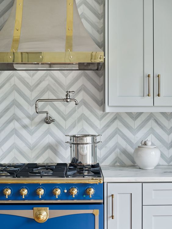 a refined and glam kitchen ith shaker cabinets, white stone countertops, a chevron tile backsplash and gold fixtures for more chic