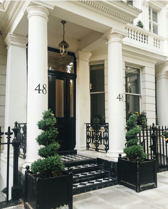 a refined black and white porch with a checked tile floor, topiaries in black pots, a lamp and a black door with glass panes