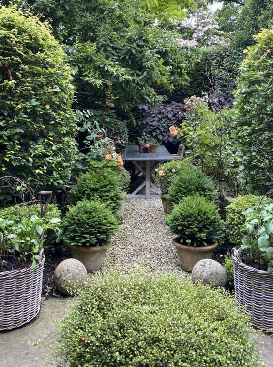 a refined shady garden with trees, greenery, potted greenery and baskets with shrubs, stone balls, a wooden dining set