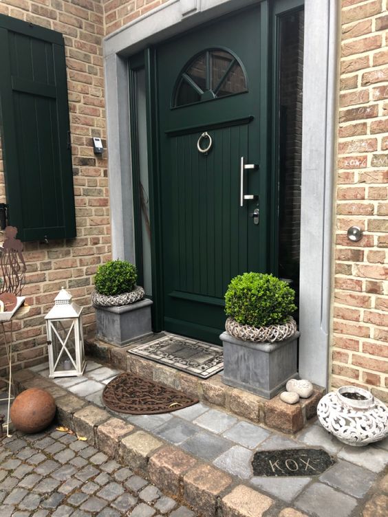 a rustic front porch with a dark green door, potted topiaries, pebbles and lanterns and some rugs is styled for the fall