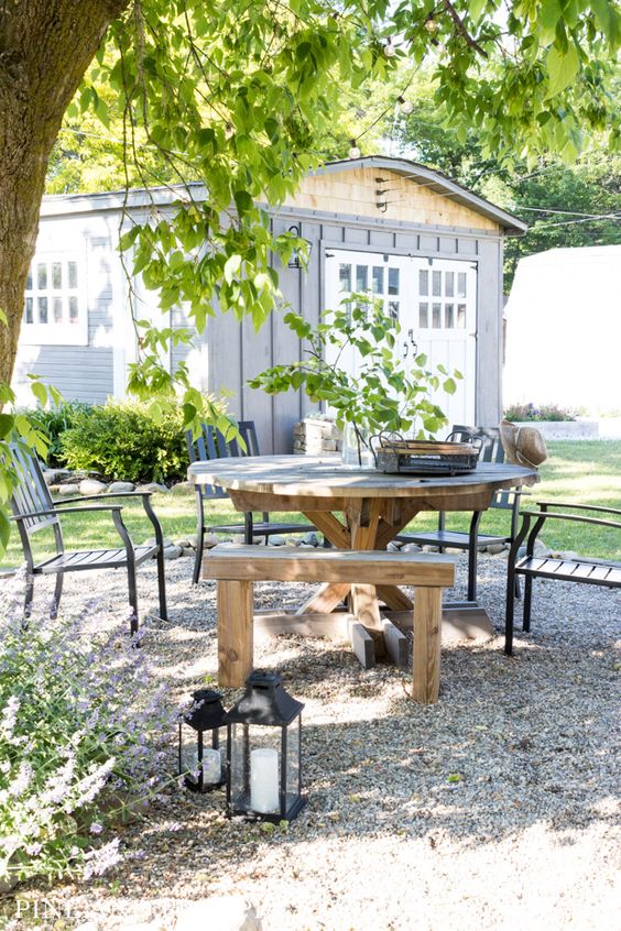 a rustic gravel patio with a round stained table and stool, black wooden chairs, candle lanterns and a tree around