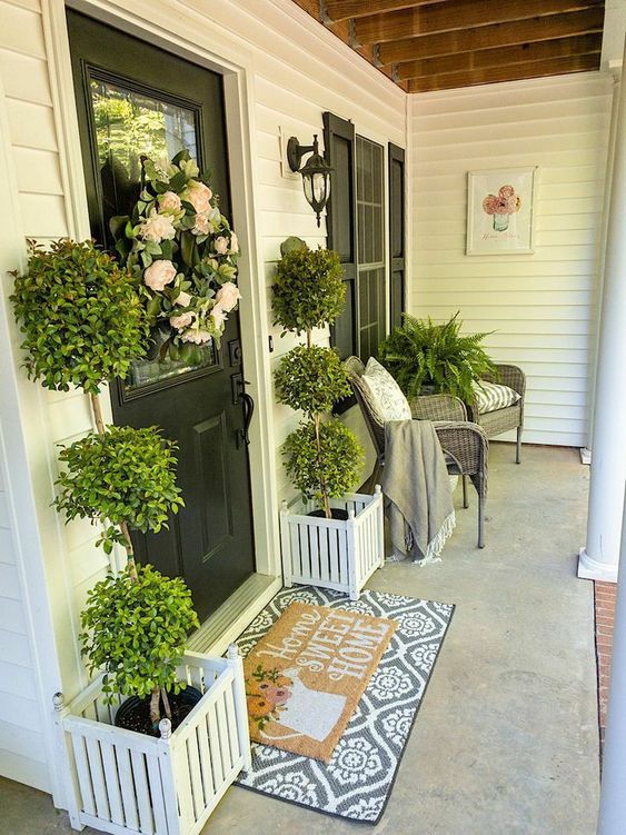 a rustic porch with wicker chairs, greenery, tall topiaries and a flower wreath plus layered rugs is amazing