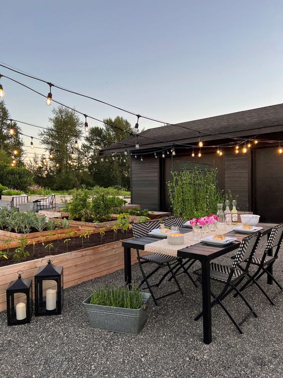 a Scandinavian gravel patio with a black table and printed chairs, potted greenery, lanterns and raised garden beds with grenery