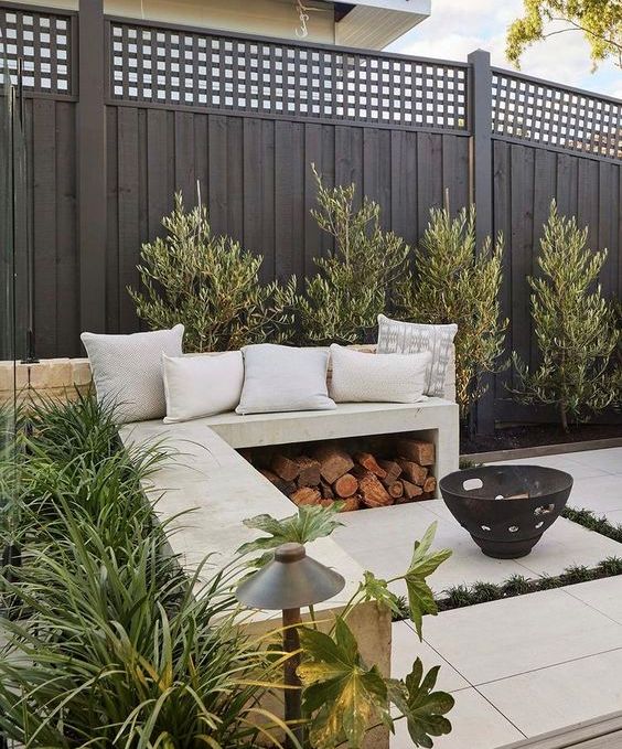 a Scandinavian nook with a black wooden fence with a lattice top, trees along it, a white concrete bench and a fire pit