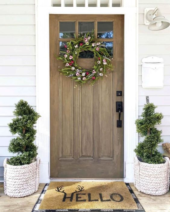 a simple rustic porch with a stained door, a wildflower wreath, topiaries framing the front door and layered rugs