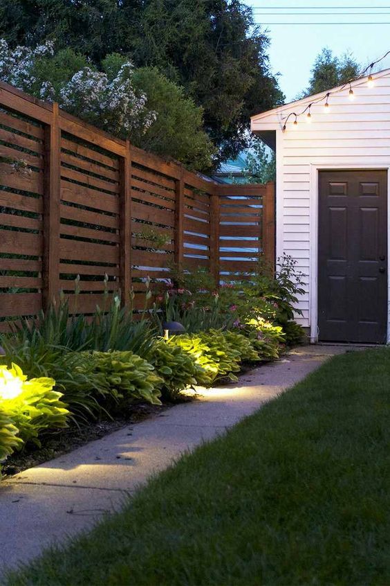 a simple stained wooden fence with greenery and lights along it are a lovely combo for a modenr outdoor space