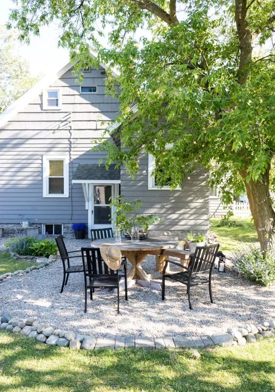 a small and cozy gravel patio with a round stained table, black chairs, rocks around a cool tree is a cool and catchy space