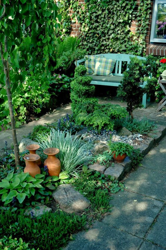 a small and cozy shady spot with greenery, a garden bed with greenery and blooms, a tree, a living wall and a blue bench with pillows