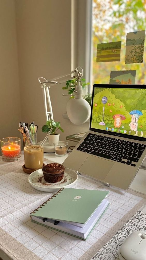 a small and cozy space with a laptop, some notebooks, some food and drinks, a table lamp and some potted greenery