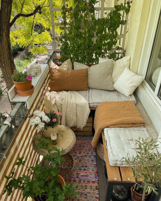 a small and cute balcony with a loveseat and a bench, pillows and blankets, side tables, greenery and blooms is amazing