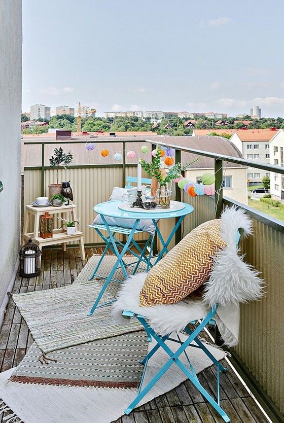 a small and cute space with blue metal furniture, layered rugs, printed pillows, lights, candle lanterns and greenery