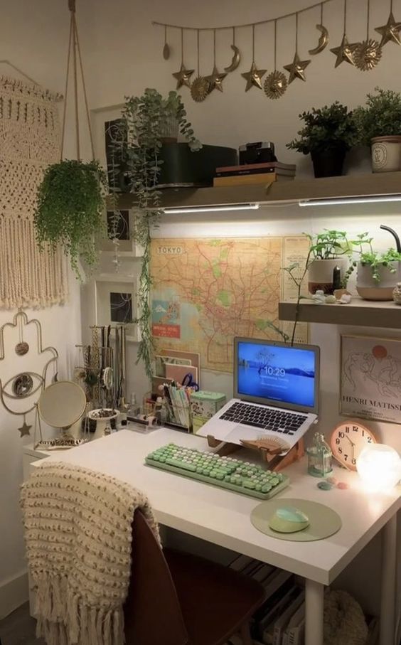 a small and lovely workspace with some shelves, potted plants, maps and artwork, a laptop and some keyboards plus boho decor on the walls