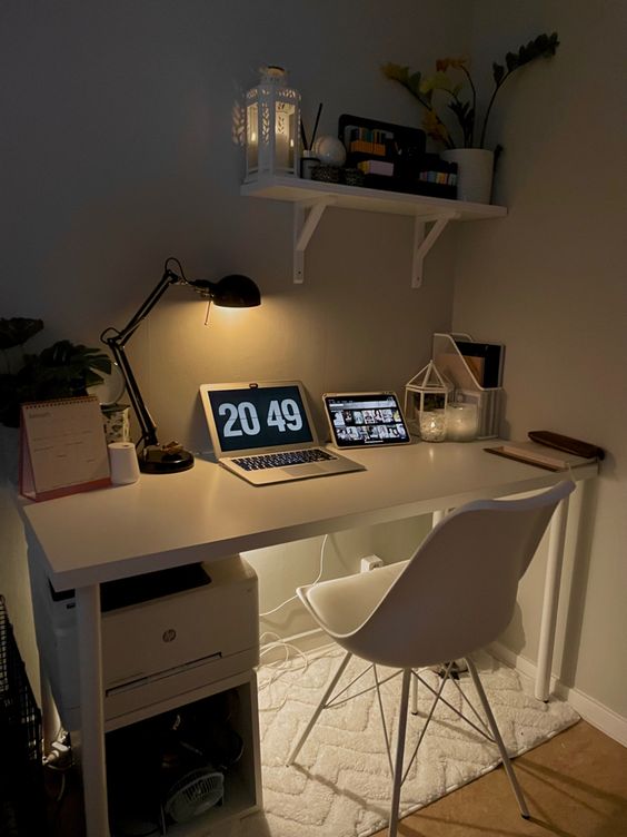 a small and simple workspace with a white desk and chair, a laptop, a table lamp and some decor, a shelf with various stuff