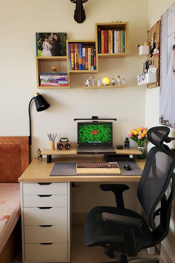 a small and well-organized workspace with bookshelves and some decor, a pegboard that holds accessories, a leptop on a stand and blooms