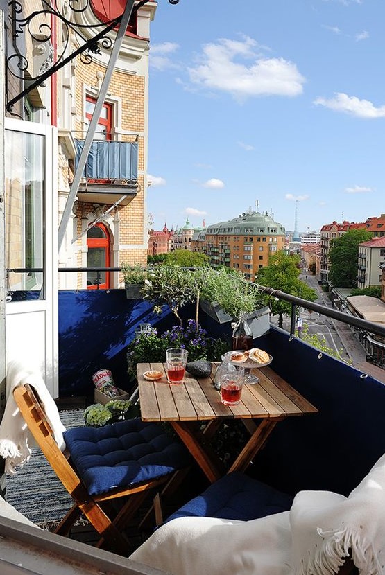 a small balcony with navy railing and upholstery, potted greenery and blooms and folding wooden furniture