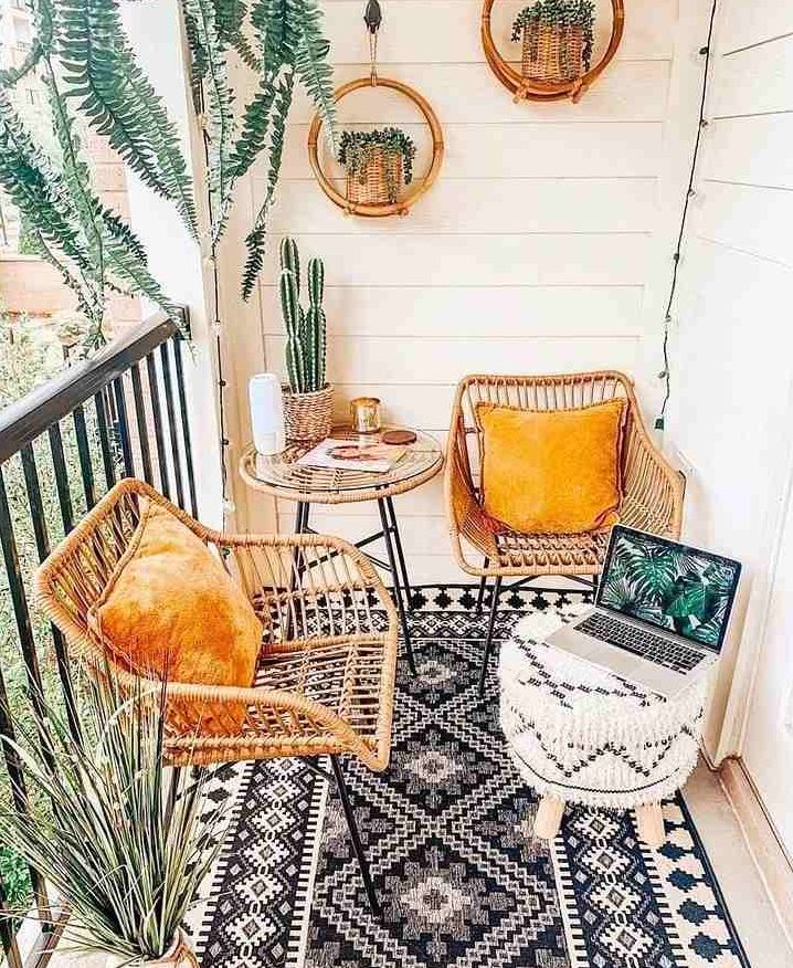 a small boho balcony with a black and white rug, rattan chairs with yellow pillows, a black and white pouf, potted plants and hanging planters